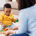 Young African American boy sits on floor facing an adult woman; they are both playing with colored blocks.