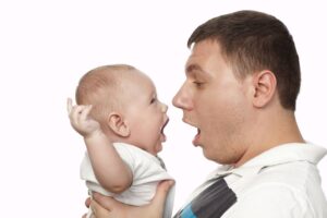 An adult male holds up a baby with its arms extended so that they are face-to-face; both have their mouths wide open.