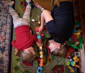 A toddler and a parent lie on the floor facing one another, playing with a train, on a multi-colored carpet. Their heads are at the bottom of the image with their bodies stretched out along the floor toward the top of the image.