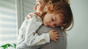 Older toddler rests head on parent’s shoulder, looks exhausted, while parent looks out a shuttered window.