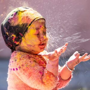 A young child with a scarf on their head, protecting their hair, is splattered with paint and closing their eyes in response to water being sprayed on their face.