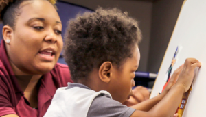 A little African-American boy works at an easel on a project while an African-American woman, seated beside him, encourages his exploration.