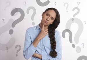 An adult woman with long dark hair, braided on the right side of her face and draped over her shoulder, stands in front of a light-colored background with floating questions marks on it.