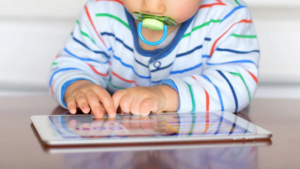 The arms and shoulders of a toddler using a pacifier and wearing a striped top are visible as the child plays with a tablet at a table.