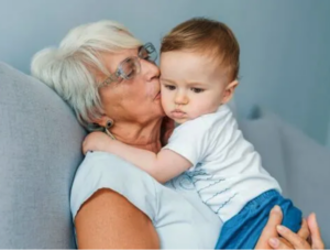 An elderly woman with short hair and glasses leans back against a light blue sofa, holding a brown-haired baby while kissing the baby’s cheek.