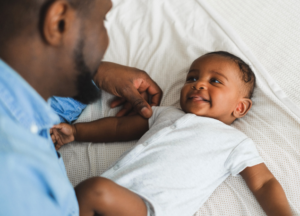 An African American father lies on the bed beside their child, looking down at the baby as the two smile at one another. The father wears a light blue shirt; the baby wears a white onesie.