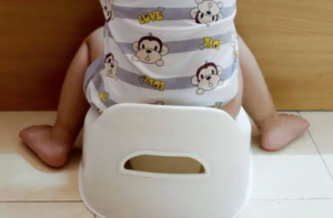 A toddler in a gray and white striped shirt with monkey faces and the word “love” on it, sits with their back to the camera on a white potty chair on the floor. The child is visible only from the shoulders down.