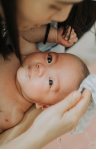 A dark-haired caregiver hovers over an infant being bathed. The infant looks up into the camera as the caregiver runs a striped washcloth over the top of the infant’s head.