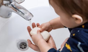 A toddler in a dark-blue shirt stands in front of a sink preparing to wash their hands under running water. An adult’s hand is holding up the bar of soap for the toddler.