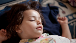 A toddler with dark curly hair lies sleeping on a dark blue background, their head turned to the side and both hands up near their head.