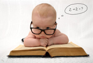 A young baby lies across an open textbook, resting their chin on their hands. The baby is wearing black horn-rimmed glasses, and we see a thought bubble beside their head with a math equation in it.