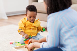 A toddler is playing on the floor with colorful toys while a caregiver sits in the foreground watching the child play.