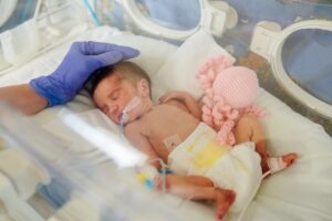 A tiny newborn sleeps in an incubator in the NICU while a caregiver wearing a purple glove reaches in and touches their head.