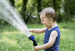 A toddler stands in a field of grass and trees, playing with a hose that is spraying water in front of the child.