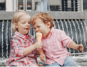 Two young children sit together on a bench in front of a fountain sharing a single ice cream cone.