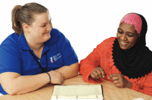 Two women, one in a blue polo shirt and the other in an orange dress and hijab, sit together at a table reviewing a document.