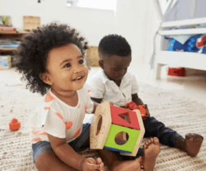 Two African-American toddlers sit on the floor together; one is playing with a wooden shape sorter while the other child plays with red shapes.