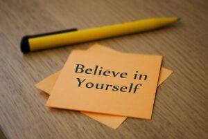 A sticky note reads "Believe in Yourself." A yellow pencil is next to it on a wooden table.