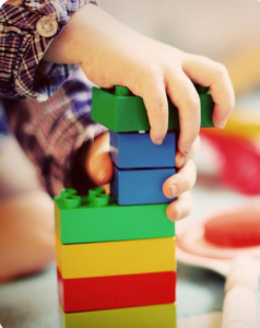 A young child uses their two hands together to stack Lego, seven bricks high.