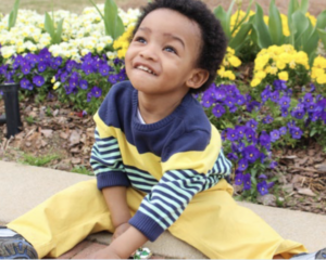 Child sitting on a sidewalk in front of purple, yellow, and white flowers.