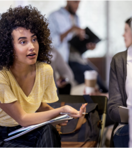 Person with curly hair holds a pen and notebook while talking in a small group meeting. 