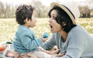 Adult and young child face each other on a blanket outdoors, practicing sounds during play. (Alternative text created using Microsoft Copilot)