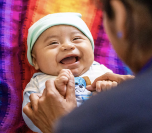 Smiling baby in a knit cap holds an adult’s hands while lying on a colorful blanket. (Alt text generated with the assist of Microsoft Copilot) 